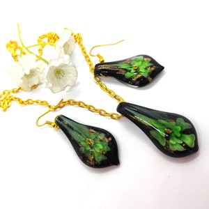 May include: A black and green floral pendant necklace and matching earrings set. The teardrop-shaped pieces feature green floral designs with gold accents. The jewelry is connected by a gold chain and set against a white background.