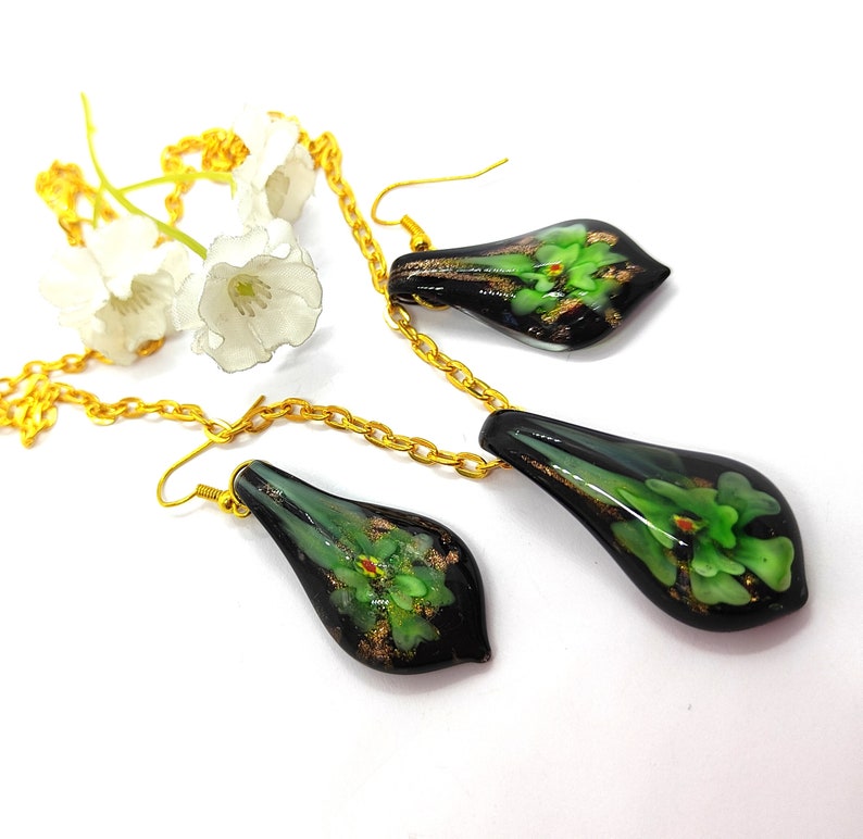 May include: A black and green floral pendant necklace and matching earrings set. The teardrop-shaped pieces feature green floral designs with gold accents. The jewelry is connected by a gold chain and set against a white background.