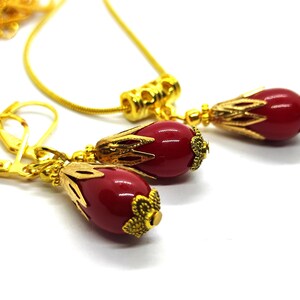 Pu&ograve; includere: Un set di gioielli color oro composto da una collana e un paio di orecchini pendenti. Ogni pezzo presenta una perla rossa liscia a forma di goccia, racchiusa in una decorativa capsula in metallo dorato con un design floreale alla base.