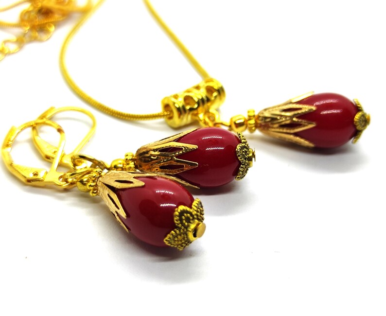 Pu&ograve; includere: Un set di gioielli color oro composto da una collana e un paio di orecchini pendenti. Ogni pezzo presenta una perla rossa liscia a forma di goccia, racchiusa in una decorativa capsula in metallo dorato con un design floreale alla base.