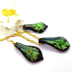 May include: A jewelry set featuring a necklace and earrings. The teardrop-shaped pendants are black with green floral designs and gold accents. The set includes a gold-colored chain and earring hooks. The background is white.