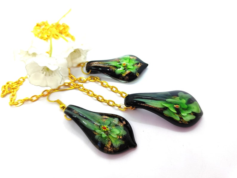 May include: A jewelry set featuring a necklace and earrings. The teardrop-shaped pendants are black with green floral designs and gold accents. The set includes a gold-colored chain and earring hooks. The background is white.