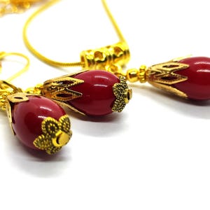 Pu&ograve; includere: Un set di gioielli composto da una collana e un paio di orecchini. Ogni pezzo presenta una perla rossa liscia di forma ovale, racchiusa in un cappuccio in metallo dorato con filigrana e una base a forma di fiore. Include una catenina dorata.