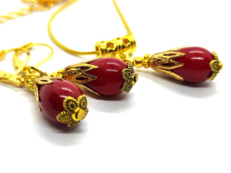 Pu&ograve; includere: Un set di gioielli composto da una collana e un paio di orecchini. Ogni pezzo presenta una perla rossa liscia di forma ovale, racchiusa in un cappuccio in metallo dorato con filigrana e una base a forma di fiore. Include una catenina dorata.