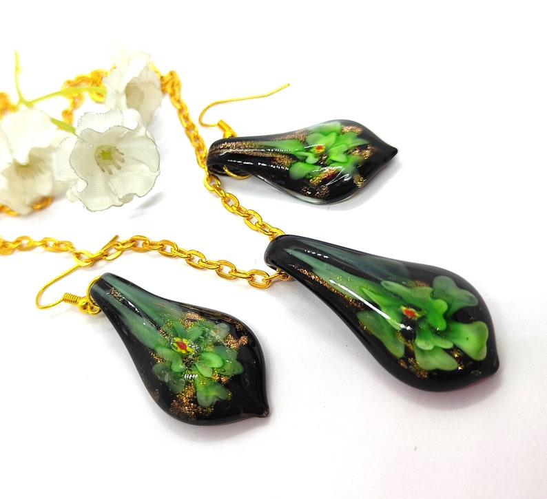 May include: A gold chain necklace and matching earrings set. The teardrop-shaped pendants are black with green floral designs and gold accents. The earrings have gold hooks. The jewelry is displayed on a white surface.