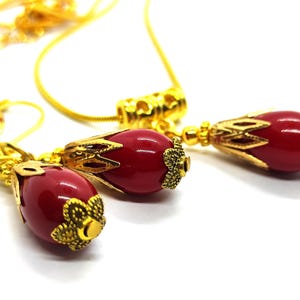 Pu&ograve; includere: Un set di gioielli composto da una collana e un paio di orecchini, ciascuno caratterizzato da una perla liscia rosso scuro a forma di goccia, racchiusa in tappi di metallo dorato con filigrana. La collana include una sottile catena dorata.
