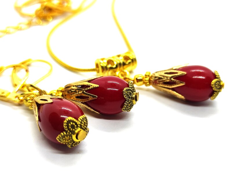 Pu&ograve; includere: Un set di gioielli composto da una collana e un paio di orecchini, ciascuno caratterizzato da una perla liscia rosso scuro a forma di goccia, racchiusa in tappi di metallo dorato con filigrana. La collana include una sottile catena dorata.