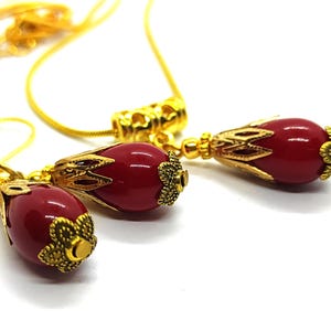 Pu&ograve; includere: Un set di gioielli composto da una collana e orecchini abbinati, ciascuno caratterizzato da una perla liscia a forma di goccia rosso intenso, racchiusa in tappi di metallo dorato con filigrana floreale, sospesi su una sottile catena dorata.
