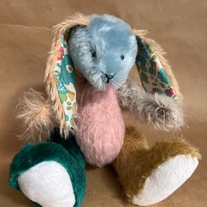 May include: A handmade plush bunny toy with a light blue head, pink body, and multi-coloured limbs. The bunny's ears feature a floral pattern. The bunny's fur is a mix of textures and colours, including brown, green, and tan.