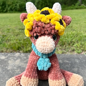 Könnte beinhalten: Ein handgefertigtes, plüschiges Kuhspielzeug, geschmückt mit einem Sonnenblumenkranz und einem blauen Schal. Die Kuh ist überwiegend braun, mit hellbraunen Hufen und einer cremefarbenen Schnauze. Der Sonnenblumenkranz hat gelbe Blütenblätter und eine dunkelbraune Mitte.