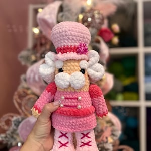 Peut inclure: Une poupée casse-noisette rose crochetée à la main. La poupée porte un chapeau rose avec une bande rouge et un nœud rose. Elle a une veste rose avec des boutons de perles et une jupe assortie. Les bottes de la poupée sont rose clair avec des lacets rouges croisés.