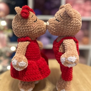 May include: Two handmade, plush teddy bear dolls. One bear wears a red dress and a red bow, the other wears red overalls. The bears are kissing. The dolls are light brown with white accents on their paws.