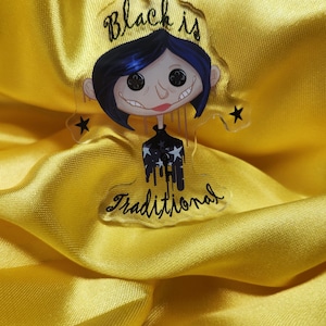 May include: A clear acrylic keychain featuring a cartoon character with blue hair, black eyes, and a black and white striped shirt. The text "Black is Traditional" is written on the keychain.