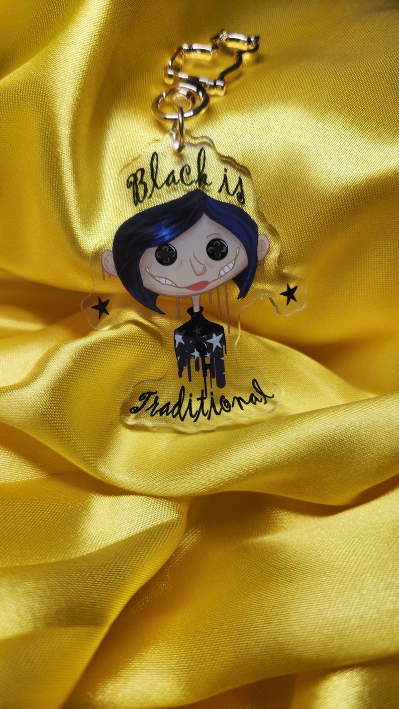 May include: A clear acrylic keychain featuring a cartoon character with blue hair, black eyes, and a black and white striped shirt. The text "Black is Traditional" is written on the keychain.