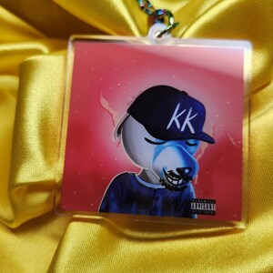 Könnte beinhalten: Ein durchsichtiger Acryl-Schlüsselanhänger mit einem Cartoon-Hund, der eine blaue und schwarze Baseballkappe mit den Buchstaben "KK" trägt. Der Hund hat ein blaues und weißes Fellmuster und einen schwarzen Bart. Der Hintergrund ist ein rosa und weißer Farbverlauf mit einem Parental Advisory-Aufkleber unten rechts.