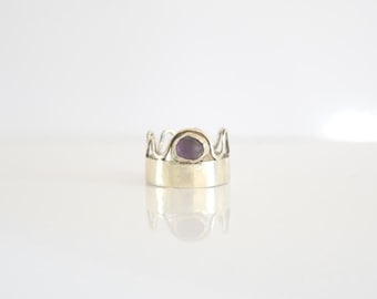 May include: A silver-toned ring with a crown-like design. The ring features a wide band and a bezel-set, oval-shaped purple gemstone. The crown design has a series of curved, wave-like shapes. The ring is displayed on a white surface.