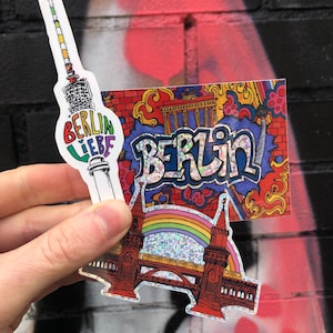 Könnte beinhalten: Zwei bunte Berliner Aufkleber. Ein Aufkleber zeigt den Berliner Fernsehturm mit den Worten "Berlin Liebe" und der andere Aufkleber zeigt eine rote Brücke mit einem Regenbogen und dem Wort "Berlin" in bunten Buchstaben.