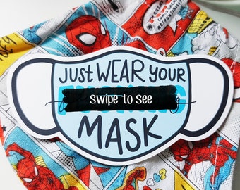 Op de afbeelding: Een blauw en wit masker met de tekst "JUST WEAR YOUR MASK" en "swipe to see" in zwarte tekst. Het masker ligt op een rode, blauwe en gele stof met een cartoon superheld ontwerp.