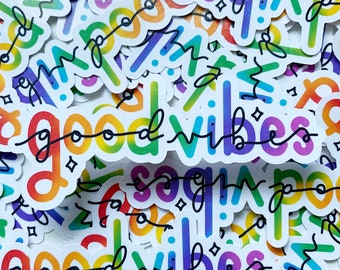 Op de afbeelding: Een close-up van een stapel regenboogkleurige stickers met de woorden "good vibes" in het zwart. De stickers zijn in een willekeurig patroon gerangschikt en zijn allemaal iets anders van formaat.