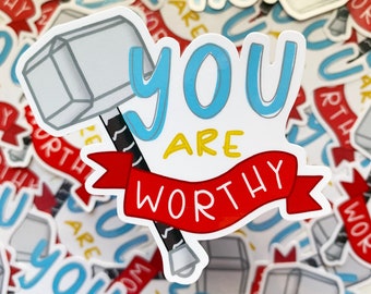 Op de afbeelding: Een witte sticker met een blauw en geel ontwerp met de tekst "You are worthy" met een rood lint en een zilveren hamer.