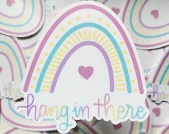 Op de afbeelding: Een sticker met een regenboogontwerp in tinten paars, blauw, geel en roze. De regenboog heeft een klein hartje in het midden en de tekst "hang in there" onderaan.
