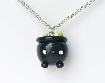 Op de afbeelding: Een ketting met een zwarte ketel en een zilveren ketting. De ketel heeft een schattig gezicht en is gevuld met groene en witte kralen.