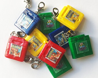 Op de afbeelding: Een set van acht kleurrijke miniatuur Game Boy-cartridges met Pokémon-spelhoezen. De cartridges zijn bevestigd aan zilveren sleutelhangers. De cartridges zijn rood, geel, blauw en groen.
