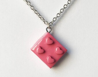 Op de afbeelding: Een zilveren ketting met een roze plastic hanger in de vorm van een Lego-steen met vier hartvormige uitsnijdingen.