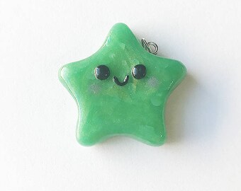 Op de afbeelding: Een groene stervormige bedel met een glinsterende afwerking en een schattig glimlachend gezicht. De bedel heeft een zilveren lus om aan een ketting of armband te bevestigen.