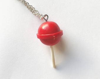 Op de afbeelding: Een zilveren ketting met een rode lolly-hanger. De lolly is op een witte stok.