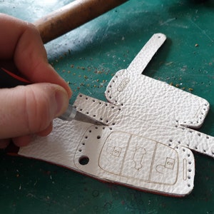 May include: A white leather key fob with a stitched border and the word "AUDI" embossed on the top. The fob is being cut with a craft knife.