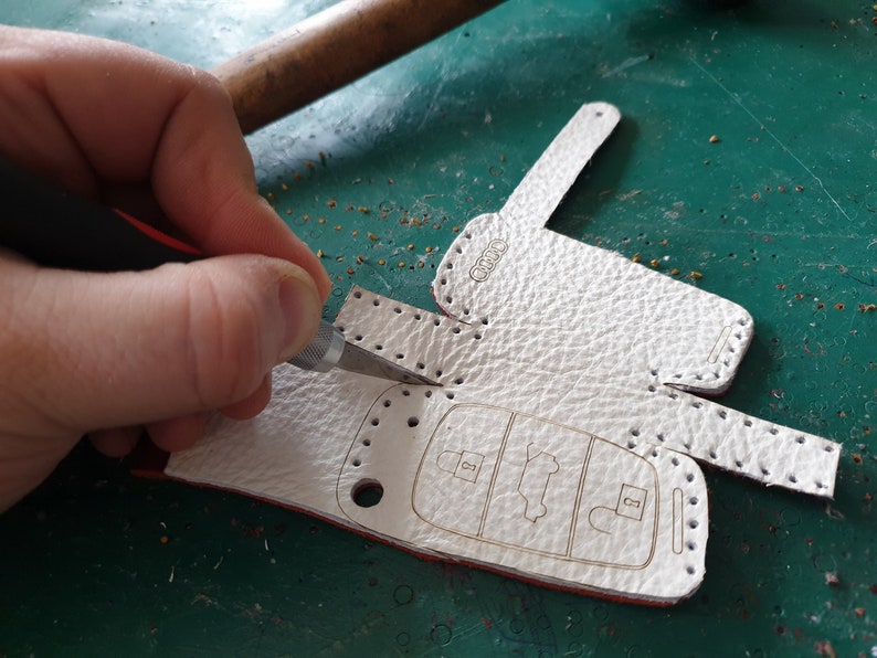 May include: A white leather key fob with a stitched border and the word "AUDI" embossed on the top. The fob is being cut with a craft knife.