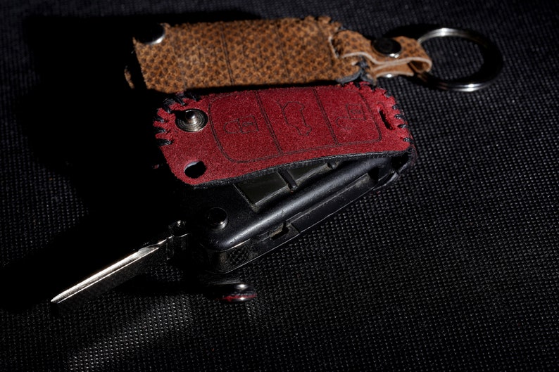 May include: A red and brown leather key fob with a silver key ring. The key fob has a black metal key attached to it.