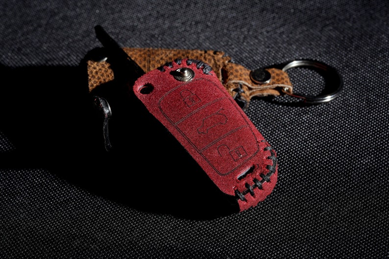 May include: A red leather key fob with stitching around the edges. The fob has a keyhole and three buttons. The fob is attached to a brown leather keychain with a silver ring.