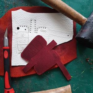 May include: A close-up of a leather craft project. A white piece of leather with a car key outline is on a red piece of leather. A red leather car key cover is on top of the red leather. A wooden mallet and a utility knife are on the table.