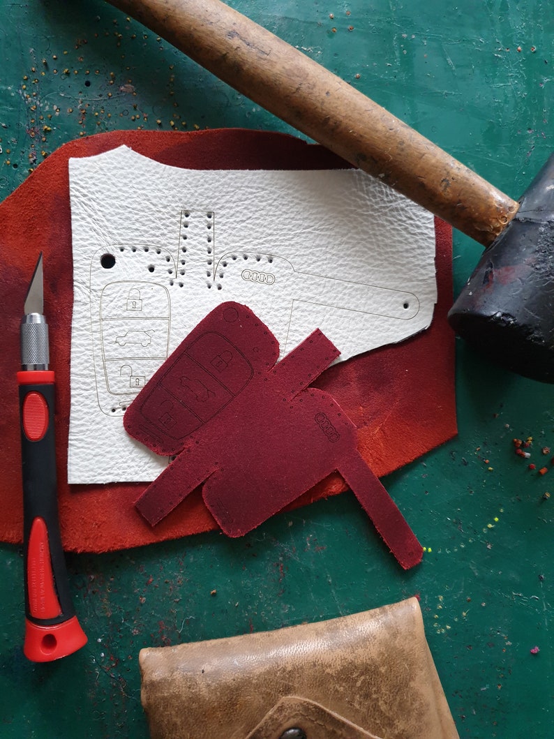 May include: A close-up of a leather craft project. A white piece of leather with a car key outline is on a red piece of leather. A red leather car key cover is on top of the red leather. A wooden mallet and a utility knife are on the table.