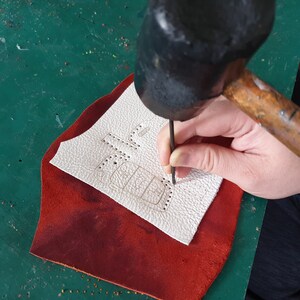 May include: A person using a hammer to punch holes in a piece of white leather. The leather has a design drawn on it and is sitting on a red piece of leather.