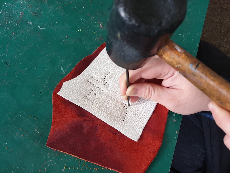 May include: A person using a hammer to punch holes in a piece of white leather. The leather has a design drawn on it and is sitting on a red piece of leather.
