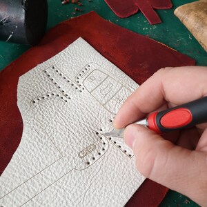 May include: A person uses a craft knife to cut a white leather piece with a design of a car key. The leather is on top of a red leather piece.