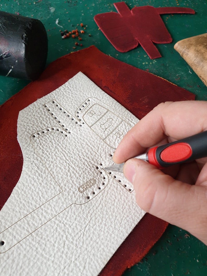 May include: A person uses a craft knife to cut a white leather piece with a design of a car key. The leather is on top of a red leather piece.
