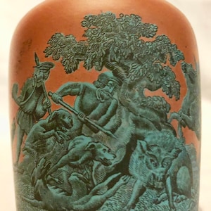 Puede incluir: Un tarro de cerámica de terracota con un diseño verde y azul que representa una escena de caza. La escena presenta a un hombre con una lanza, un perro y un jabalí.