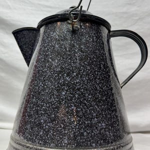 Puede incluir: Cafetera vintage de esmalte moteado. La cafetera es negra con motas blancas y azules. Tiene un asa negra y una tapa con bisagras. El pico es triangular y el asa es de alambre metálico curvado con un mango de madera negro.