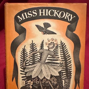 May include: Vintage book cover for "Miss Hickory" by Carolyn Sherwin Bailey. The cover features a whimsical illustration of a doll-like figure in a leaf dress, surrounded by ferns and flowers, with a bird overhead. The title is in bold white letters.