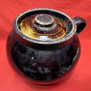 Puede incluir: Una olla redonda de cerámica marrón oscuro con tapa y asa. La olla tiene un acabado brillante con tapa y borde marrón claro. La tapa tiene un pequeño pomo redondo. La olla está sobre una superficie roja.