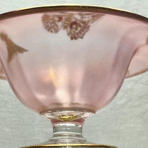 May include: A pink glass bowl with a gold rim and a decorative gold design. The bowl has a pedestal base and a wide, shallow shape. The glass has a frosted appearance.
