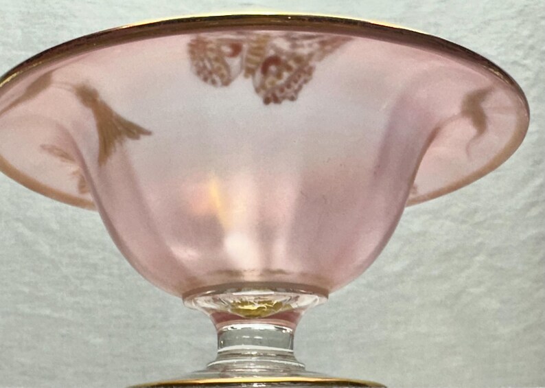 May include: A pink glass bowl with a gold rim and a decorative gold design. The bowl has a pedestal base and a wide, shallow shape. The glass has a frosted appearance.