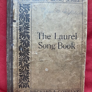 May include: An antique book titled "The Laurel Song Book" from the Laurel Music Series. The cover is a faded tan color with black decorative elements and text. The publisher's name, C. C. Birchard & Company, is printed at the bottom.