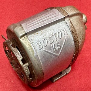 May include: Vintage silver-toned pencil sharpener with the text "BOSTON KS" embossed on the side. The sharpener has a crank handle and a circular blade housing. The metal shows signs of age and use, with some rust.