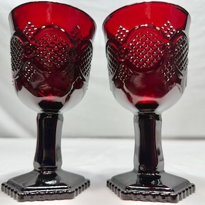 May include: A pair of deep red glass goblets featuring a textured diamond pattern on the upper bowl, a smooth cylindrical stem, and a square base with a beaded edge. The glass is translucent and reflects light across its faceted surface.
