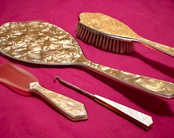 May include: A vintage vanity set with a large hand mirror, a hair brush, a nail buffer, and a cuticle pusher. The set features a gold and cream-colored, textured design with silver trim. The items are arranged on a deep pink surface.